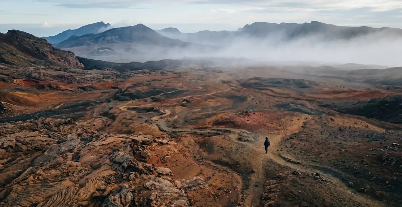 Paysage volcanique rouge et noir de la Plaine des Sables avec un randonneur solitaire au loin, entre brume matinale et ciel bleu